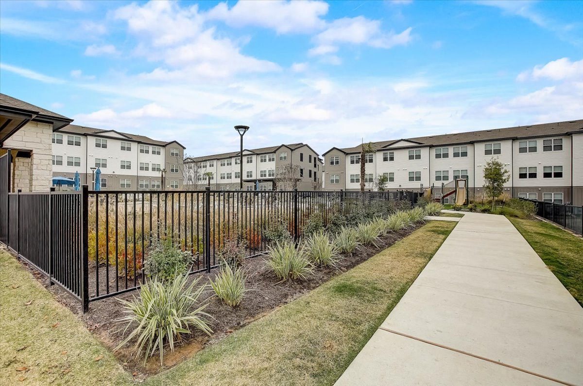 Landscaped path beside a fenced green space, with ornamental plantings and apartment buildings beyond.